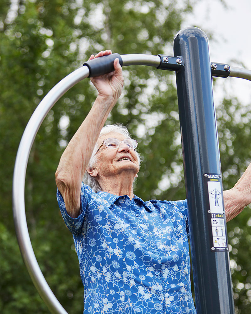 An elderly woman exercises on an arm swirl for flexibility at an outdoor fitness area.